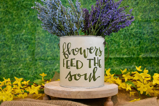 A white bucket with green lettering that says, Flowers feed the soul. There are purple and blue faux flowers in the bucket.