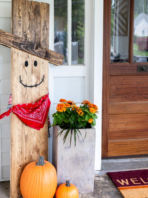 a front porch scene with ahandmade, wood scarecrow, pumpkins, and orange flowers next to a door