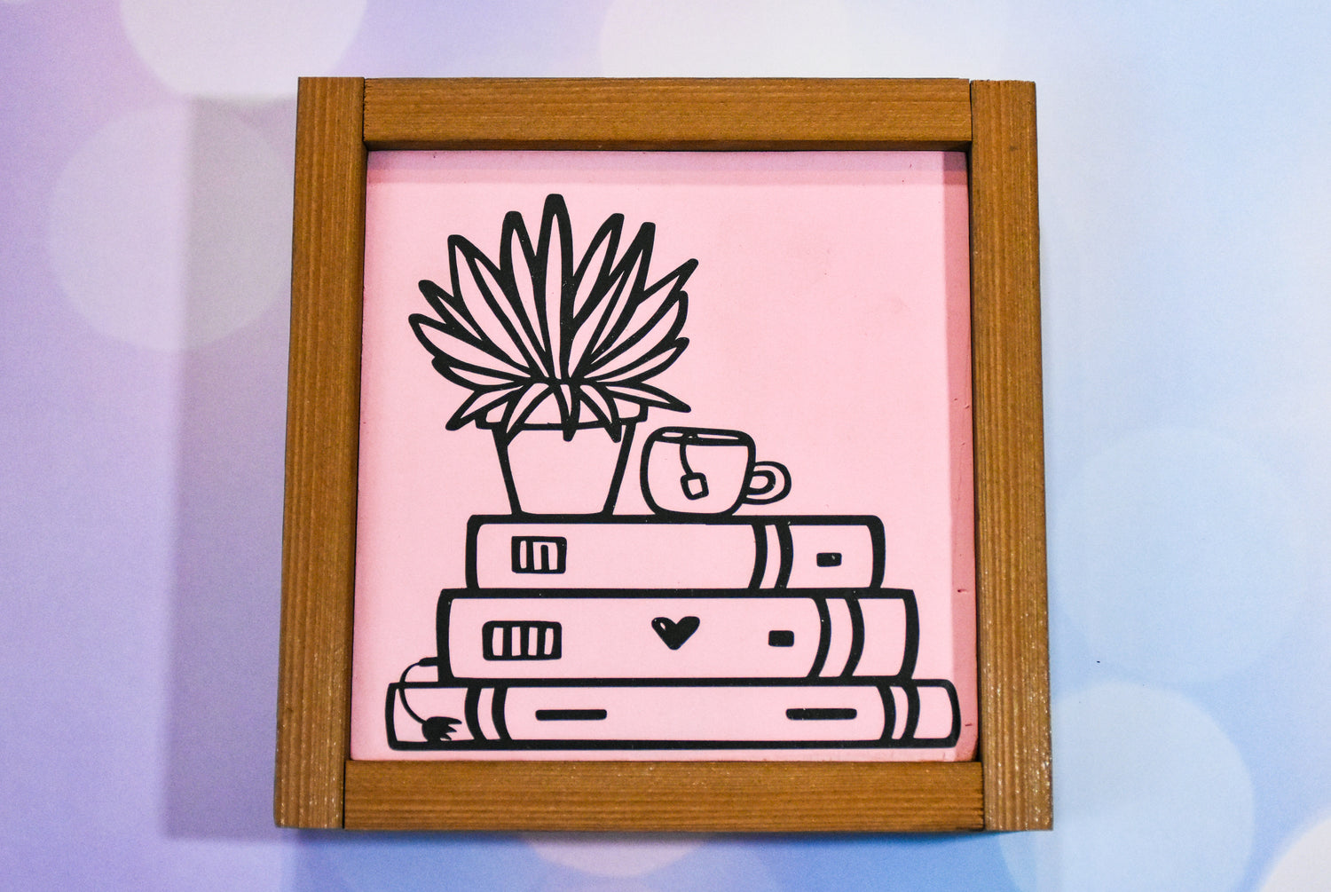 A framed wood square, painted pink, with black vinyl stack of books, a plant, and a cup of tea.